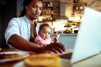 Man working on his laptop at breakfast, with his baby on his lap.